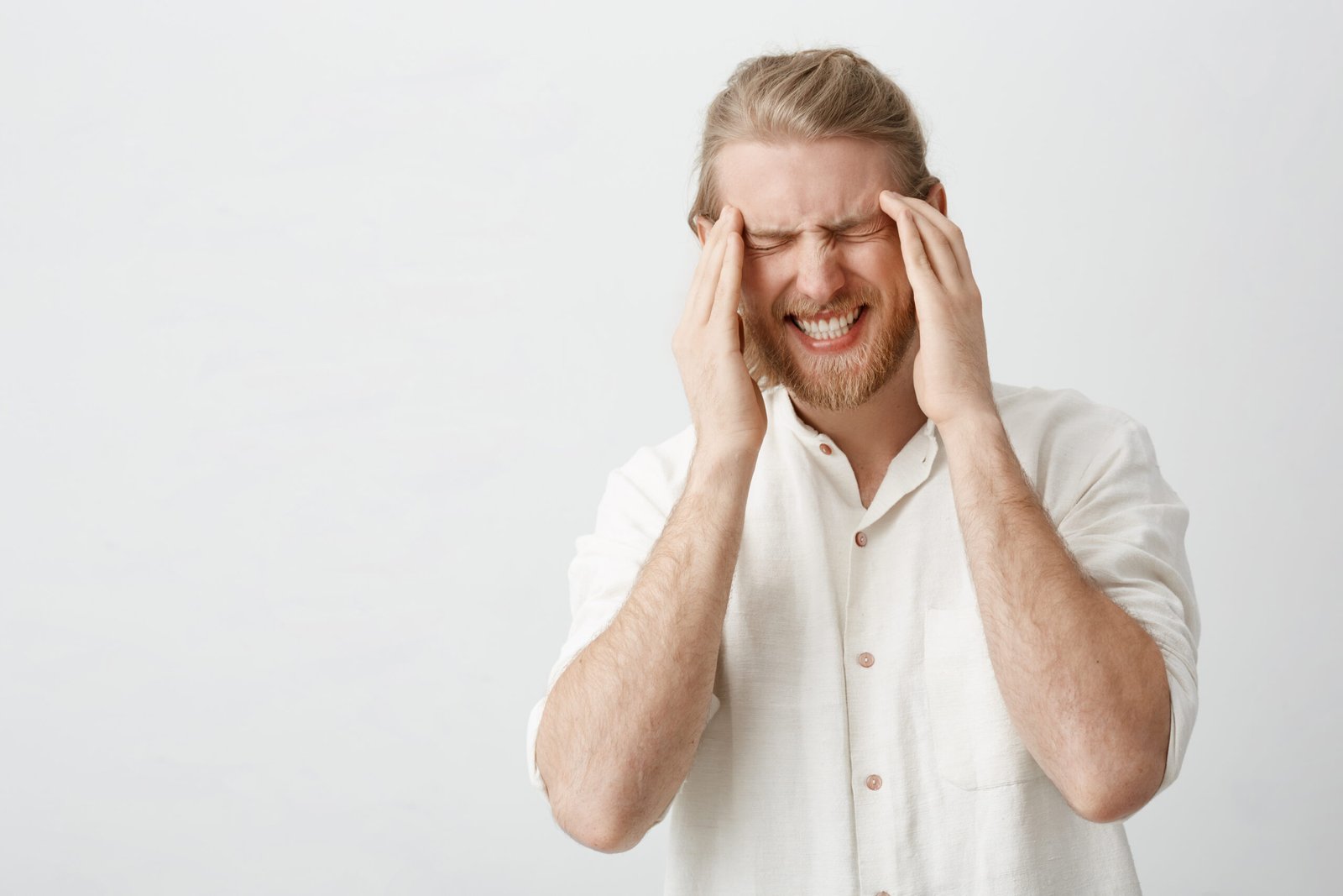 Indoor portrait of caucasian blond man with beard suffering from migraine or headache, holding fingers on temples, frowning and squeezing teeth from pain, standing over gray background. Copy space