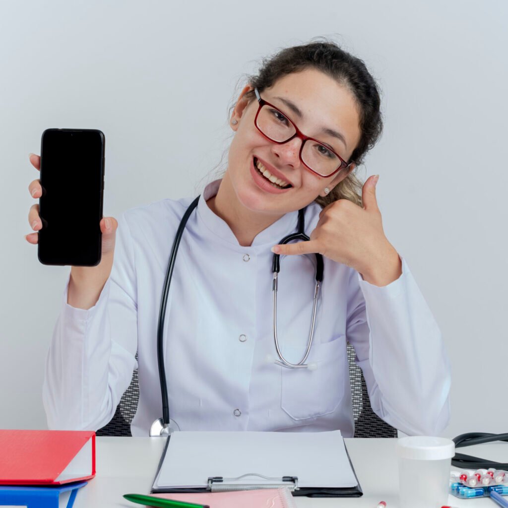 joyful young female doctor wearing medical robe and stethoscope and glasses sitting at desk with medical tools showing mobile phone looking at camera doing call gesture isolated on white background